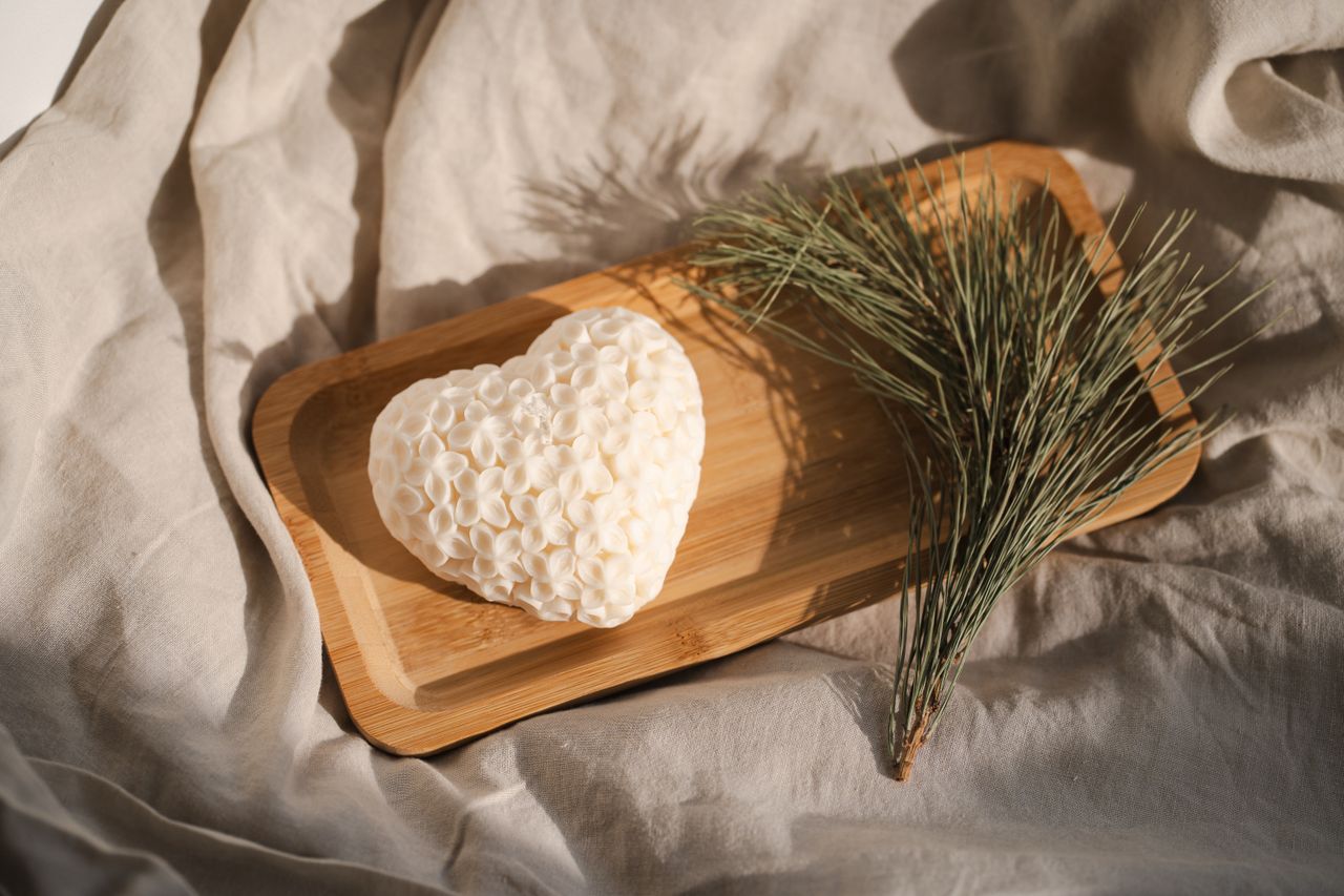 A heart-shaped white textured soap bar resting on a rectangular wooden tray, styled with a sprig of pine needles on draped beige fabric.