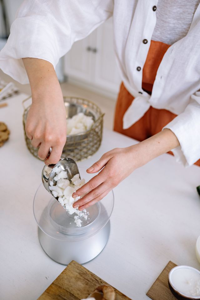 A person in a white shirt and rust-colored apron scoops white wax or soap chunks into a clear container on a white countertop in a bright kitchen setting.
